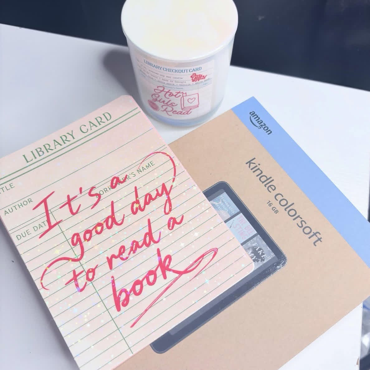 A Kindle Colorsoft Fire book token card with a clear window showing a coin inside, placed next to a 'library card' note that says 'It's a good day to read a book' in pink script, with a white candle in the background on a white surface.