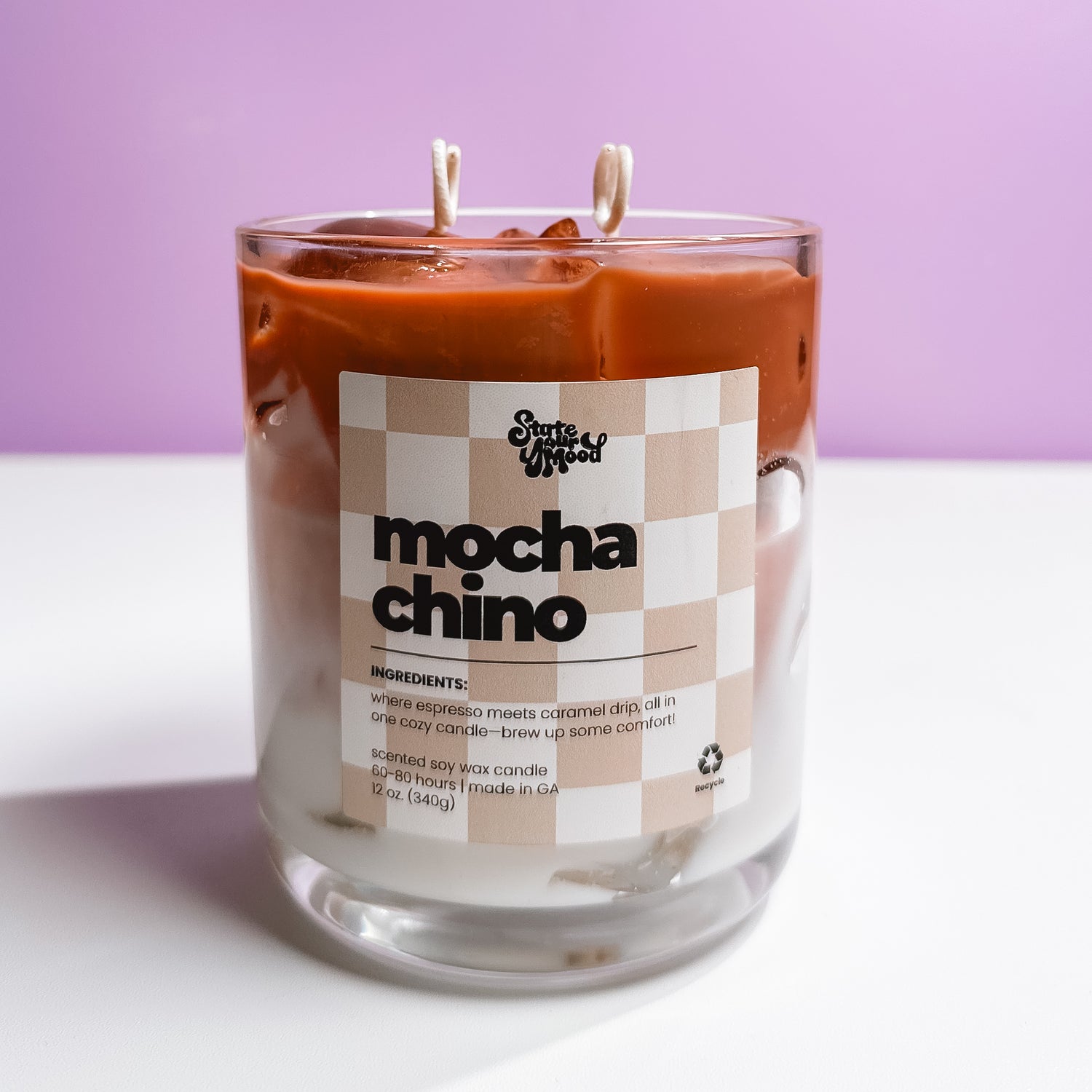 Iced coffee-inspired soy wax candle with wax ice cubes, labeled "mochachino" in a clear glass jar, featuring creamy coffee swirl layers and dual wicks. Hand-poured scented candle by State Your Mood, photographed against a pastel purple background.