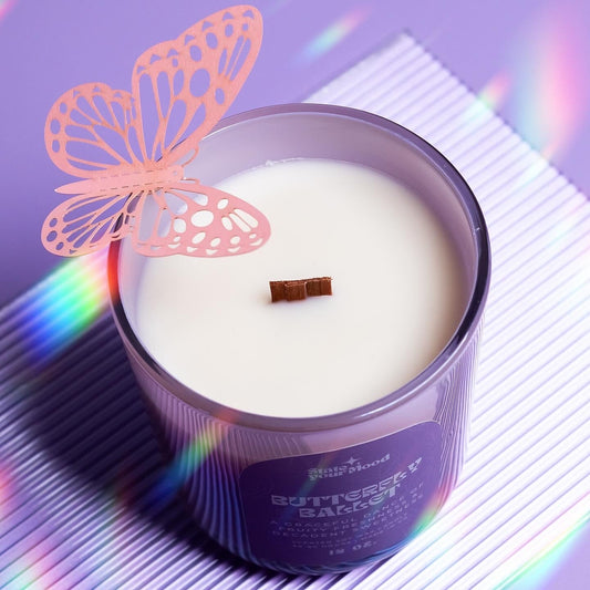 A top-down view of the 'Butterfly Ballet' candle by State Your Mood. The candle is in an iridescent purple glass jar with a wooden wick in the center of the white wax. A decorative orange butterfly is attached to the rim of the jar. The background features soft, rainbow-like reflections on a white, striped surface, creating a dreamy, whimsical vibe.