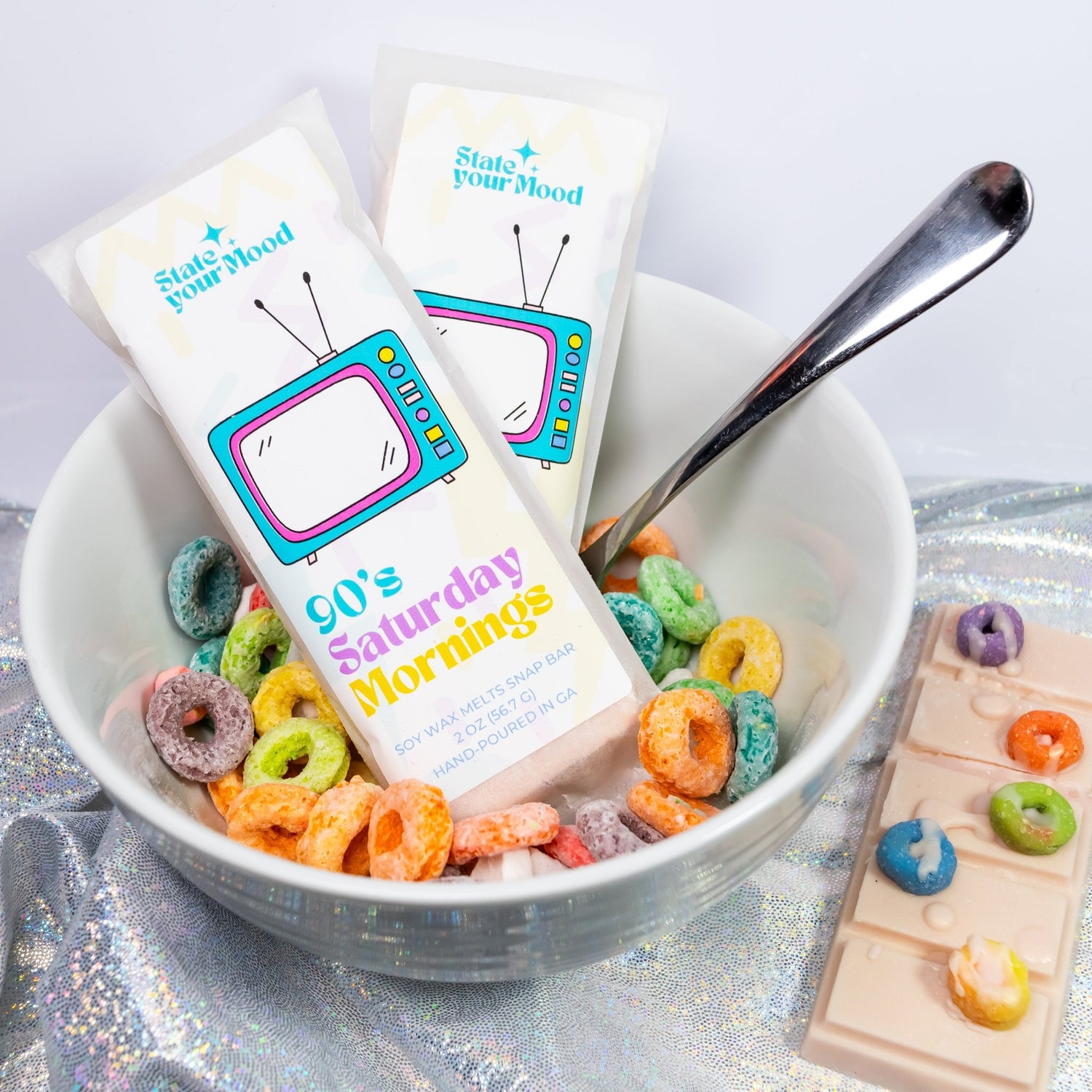 A nostalgic setup featuring the '90's Saturday Mornings' wax melt snap bar by State Your Mood. The packaging has a retro TV design with bold, colorful text. Two snap bars are placed in a bowl filled with fruit-flavored cereal, with a spoon resting inside. To the right, a wax melt snap bar decorated with multicolored cereal-shaped pieces is displayed on a shimmering surface, enhancing the playful and retro theme.