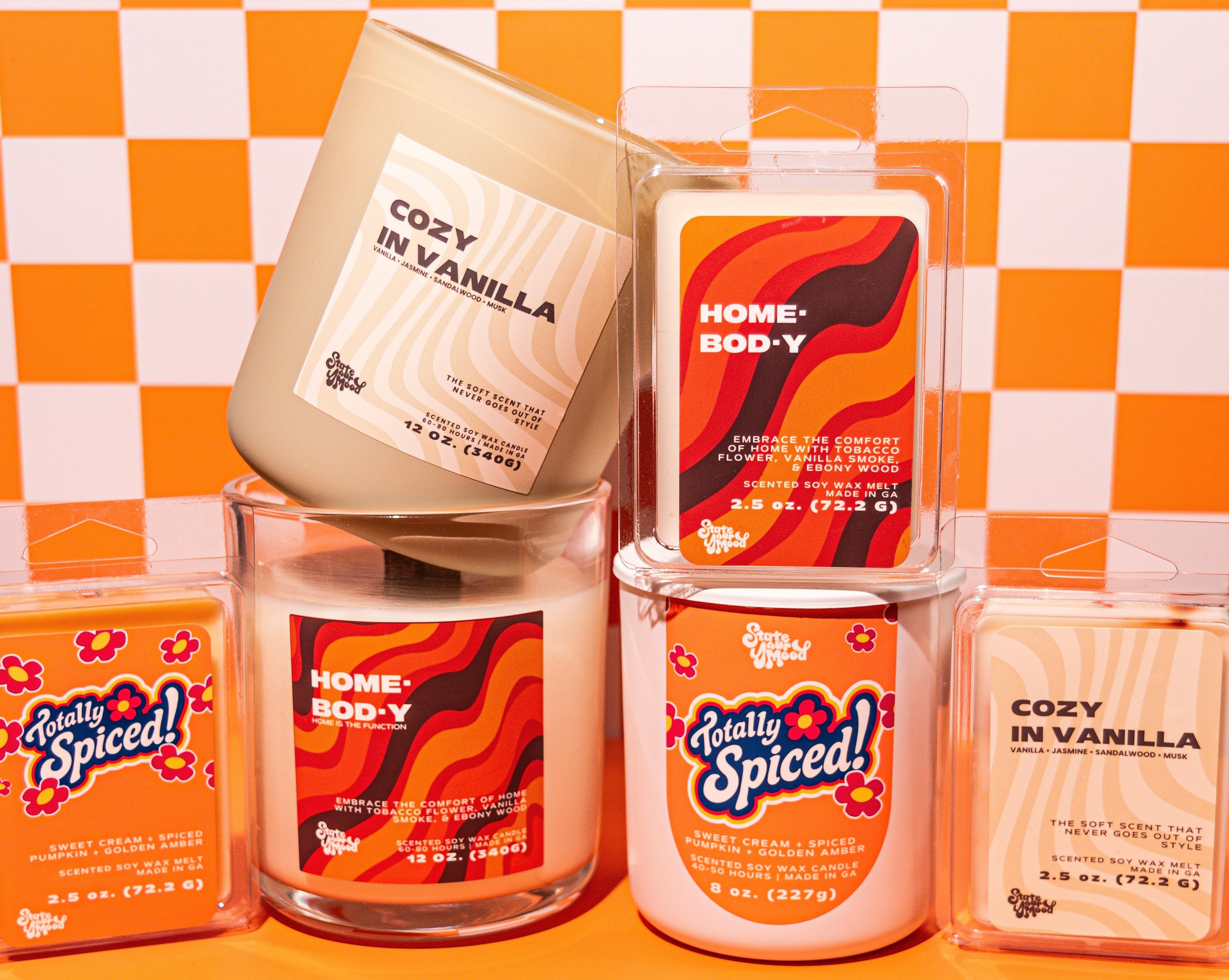 A group of scented candles and wax melts from the brand Pretty Spiced! arranged on a bright orange surface against a white and orange checkered background. The products include a large cream-colored jar labeled 'Cozy in Vanilla' and several smaller wax melts in clear packaging with orange and red wavy labels.