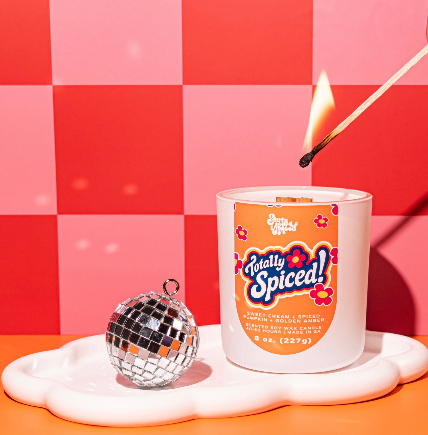 A white jar candle labeled 'Totally Spiced!' sits on a white cloud-shaped tray beside a small mirrored ornament, with a lit match hovering above the wax. The background is a pink and red checkered wall with an orange surface.