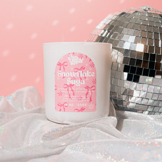 This image features the Snowflake Suga Candle, presented in a charming matte white vessel with a minimalist design. The candle, housed in a shimmering pink and white bell bottom, adds a festive touch to the serene holiday-themed backdrop.